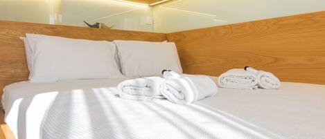 Una cama bien hecha con ropa de cama y toallas blancas, cabecera de madera e iluminación empotrada.
