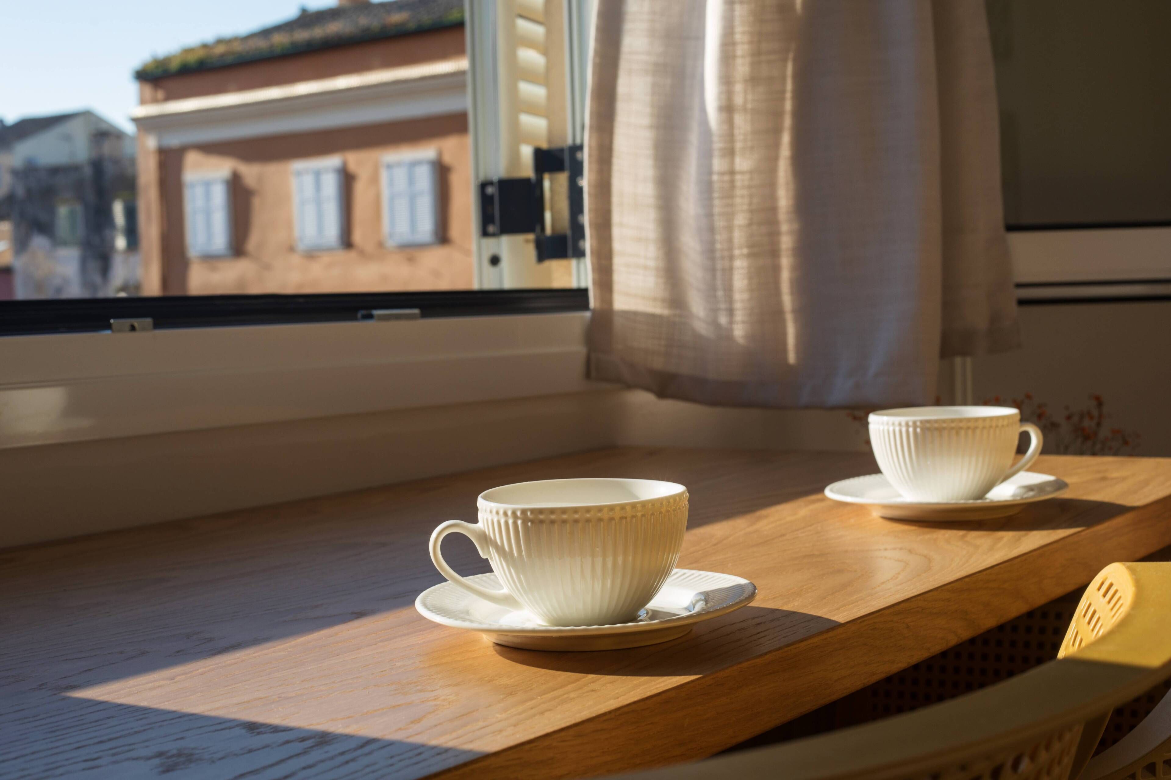 A window with a view of a building, a wooden table with two white cups and saucers, and a yellow chair.