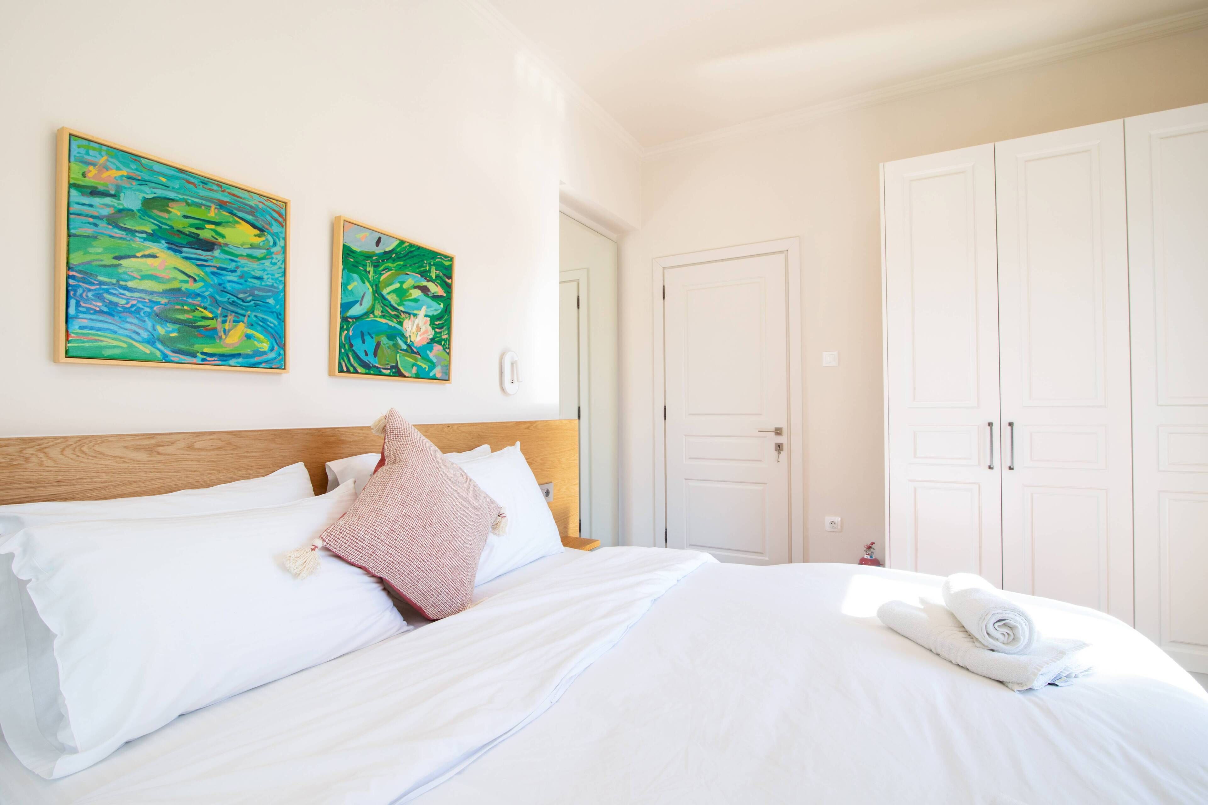 Ein Schlafzimmer mit einem hölzernen Kopfteil, weißer Bettwäsche, zwei Bildern an der Wand und einem Kleiderschrank.