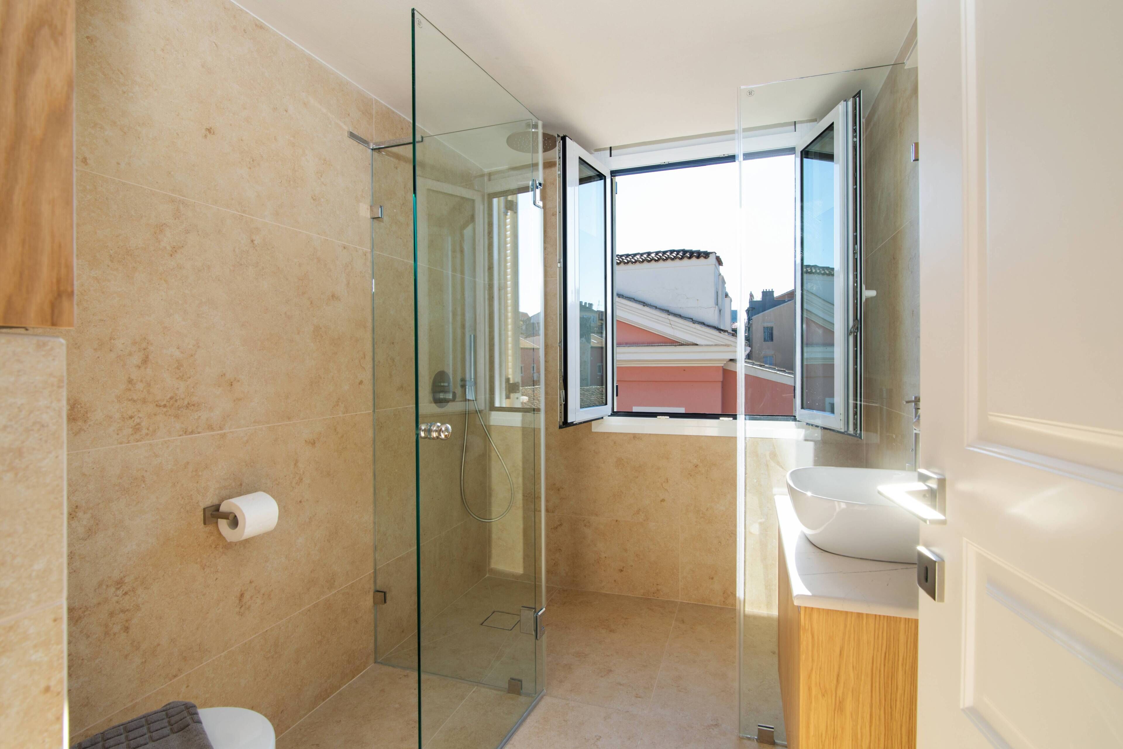 A modern bathroom with a glass shower enclosure, a toilet, a sink, and a window offering a view of buildings outside.