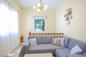 Uma sala de estar com um sofá cinzento, uma cabeceira de madeira, um candelabro, uma janela com vista para árvores e um extintor de incêndio.
