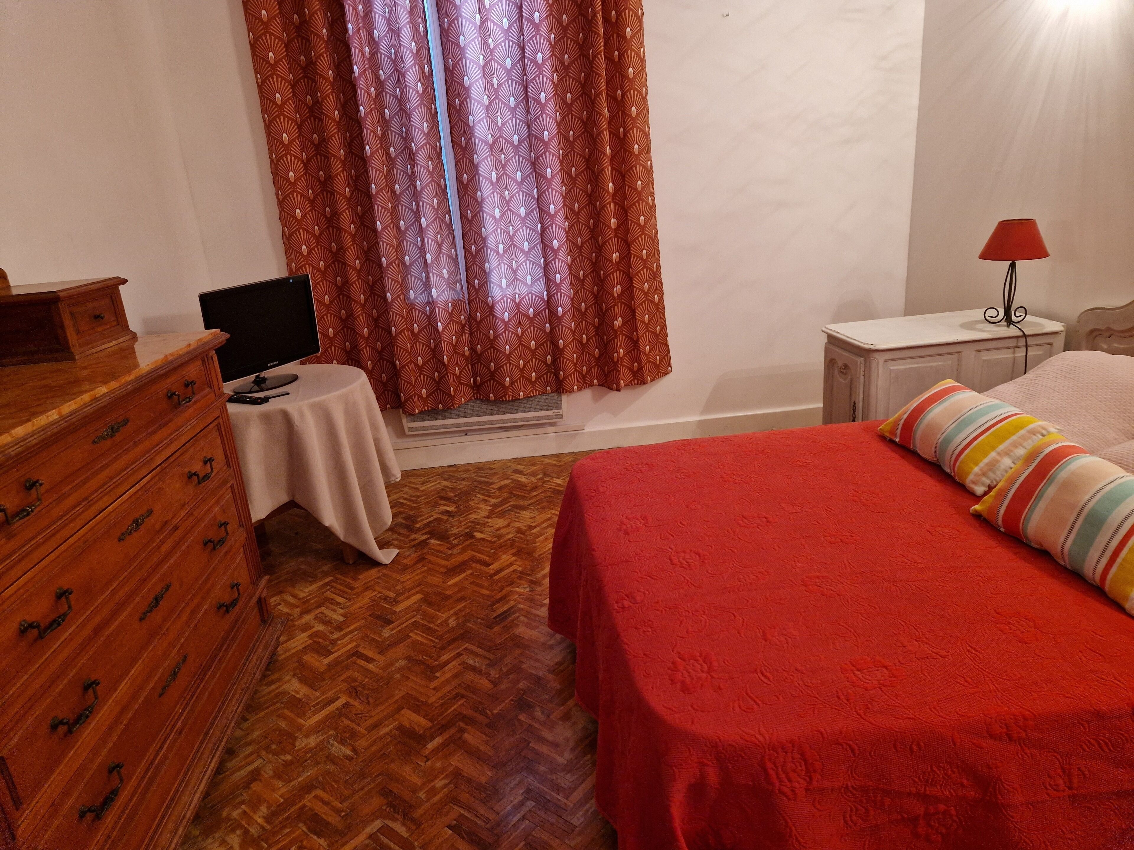 A bedroom with a wooden dresser, a small table with a lamp, a bed with a red bedspread, and a window with patterned curtains.