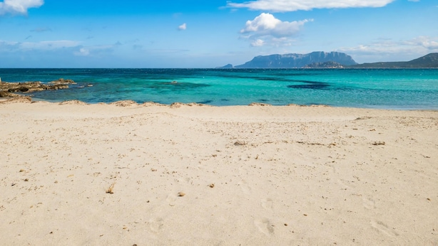 Una playa con agua turquesa cristalina y montañas lejanas bajo un cielo azul.