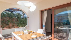 Mesa de madera con sillas blancas en un patio exterior, puerta de cristal que da a un balcón y vista a un jardín.