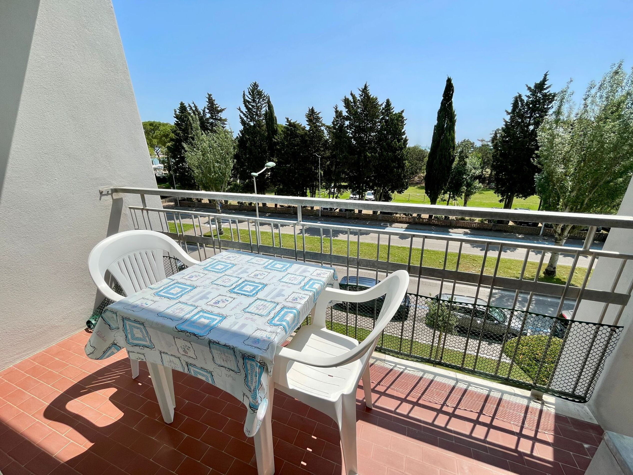 A balcony with a table and chairs, a view of a street and trees, and a metal railing.