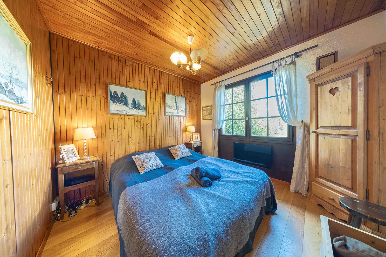 A wooden-paneled room with a bed, a window with curtains, a wooden cabinet, and a wall-mounted lamp.