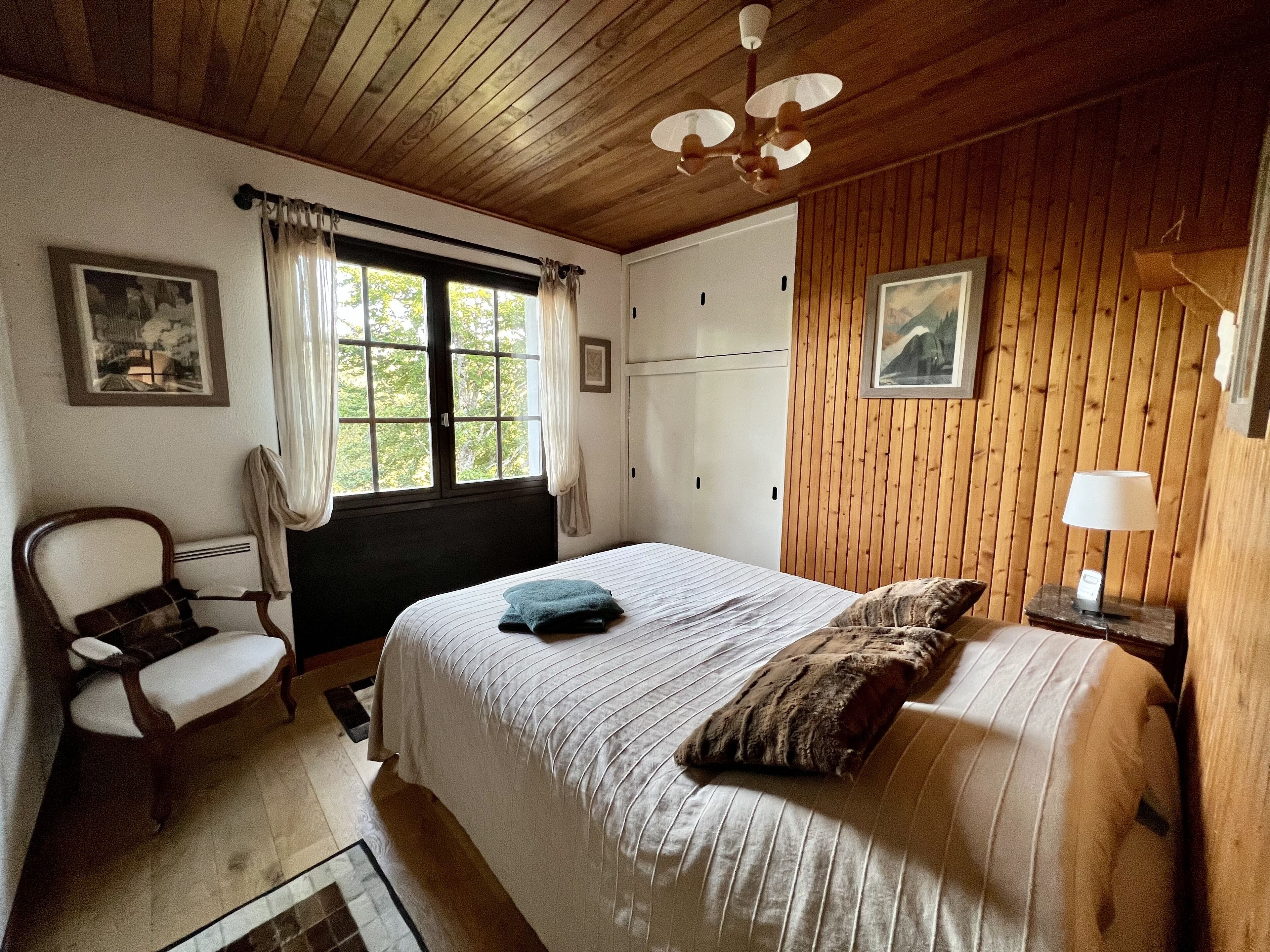 Un dormitorio con techo de madera, una cama con ropa de cama blanca, una silla, un ventanal con cortinas y un cuadro en la pared.
