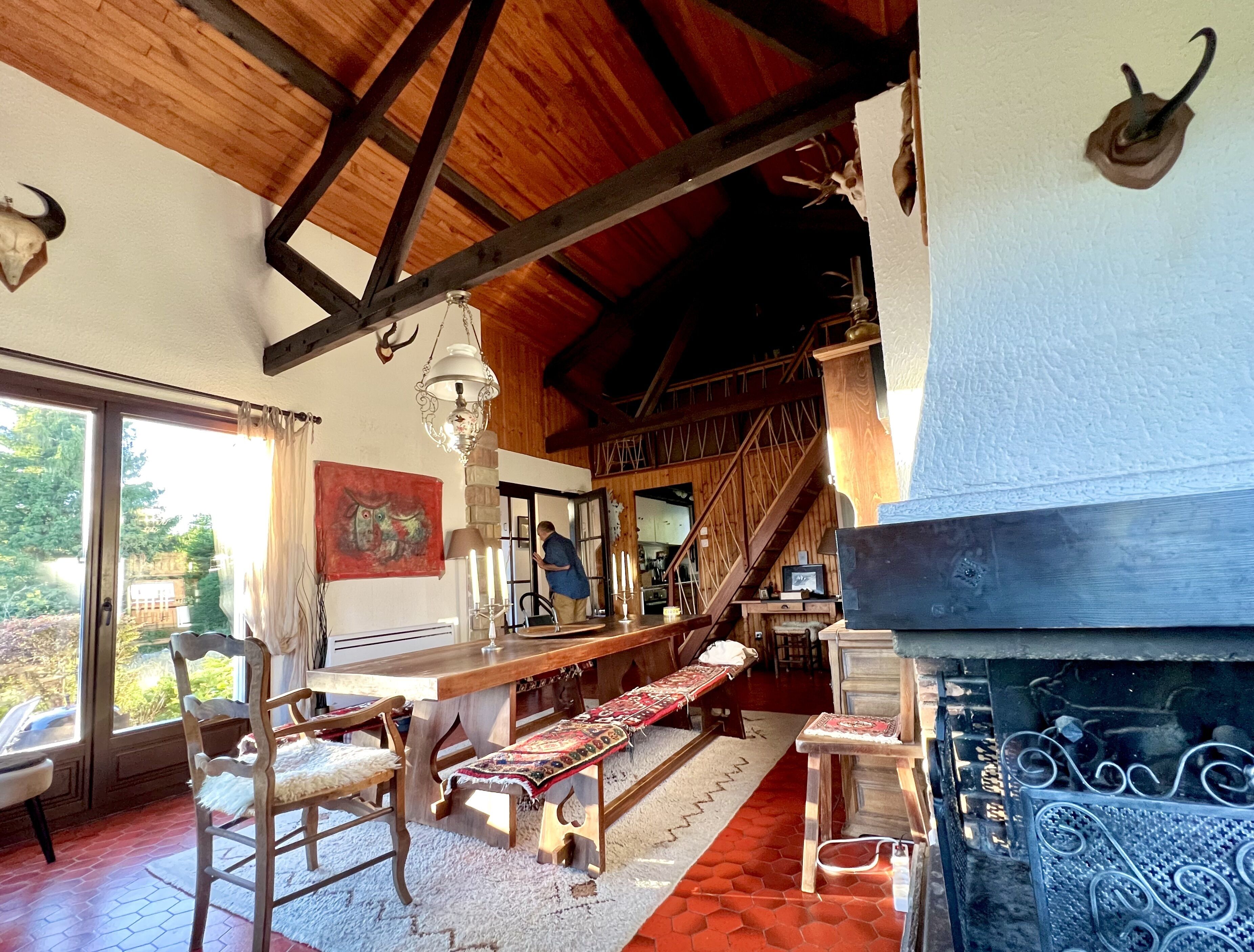A rustic interior with wooden beams, a dining area with benches, a fireplace, and a view of the outdoors.