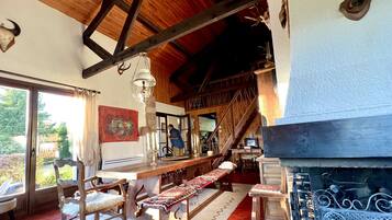 A rustic interior with wooden beams, a dining area with benches, a fireplace, and a view of the outdoors.