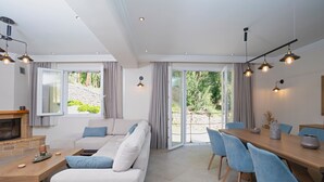 Uma sala de estar moderna com sofá, mesa de centro e área de jantar com cadeiras. Há uma lareira, janelas amplas e vista para o verde lá fora.