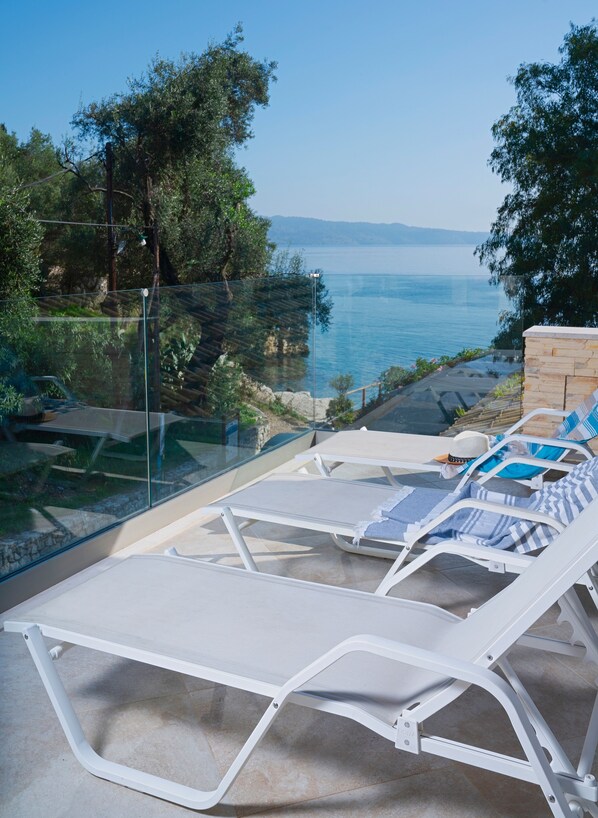 A terrace with white lounge chairs, a glass balustrade, and a view of the sea.
