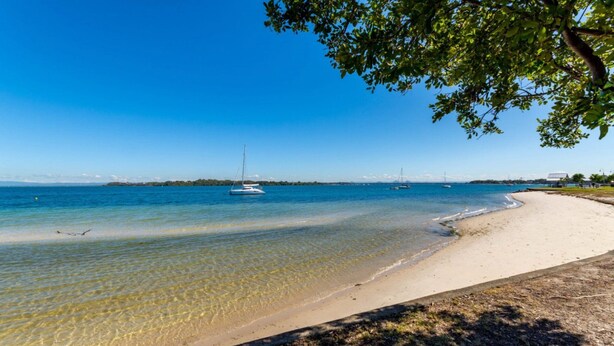 A beach with clear shallow waters, a sandy shore, and sailboats anchored in the distance.