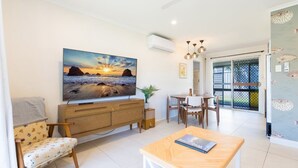 A modern living room with a large TV, wooden furniture, a dining area, and a view of the outdoors.