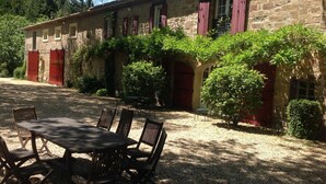 A stone building with a gravel courtyard, a table with chairs, and red shutters.