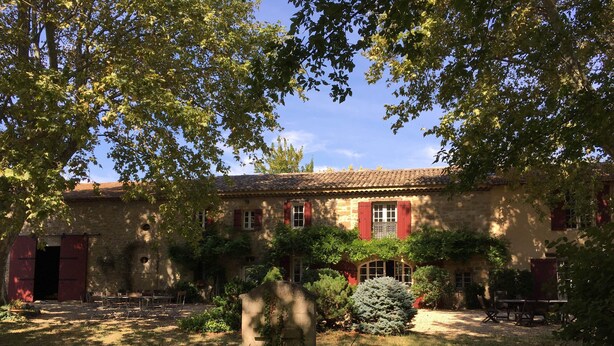 A rustic stone building with red shutters, surrounded by lush greenery and a well-maintained garden.
