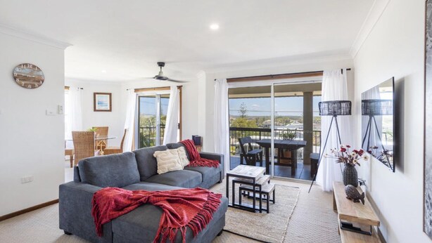 A modern living room with a grey sofa, a red throw blanket, and a balcony with a table and chairs.