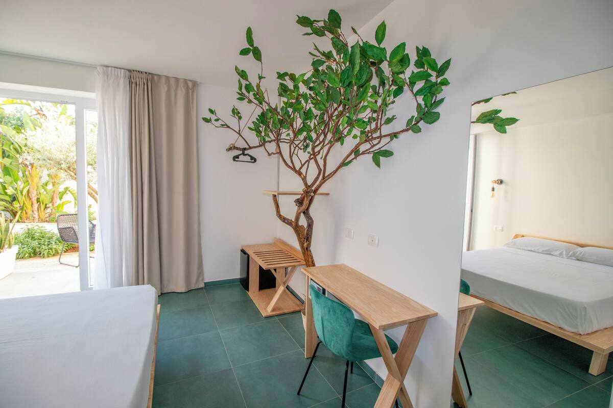 A room with a bed, a desk, a chair, a small pet bed, and a large artificial tree.