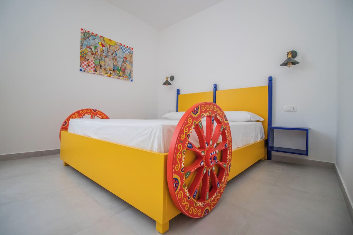 A bed with a yellow frame and a large red wheel, a small bedside table, and a wall-mounted light fixture.