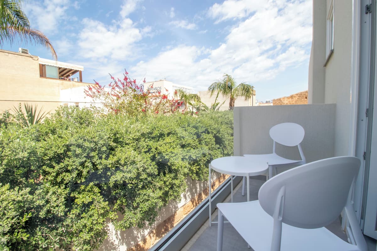 A balcony with white chairs and a table, overlooking a garden with various plants and a building in the background.