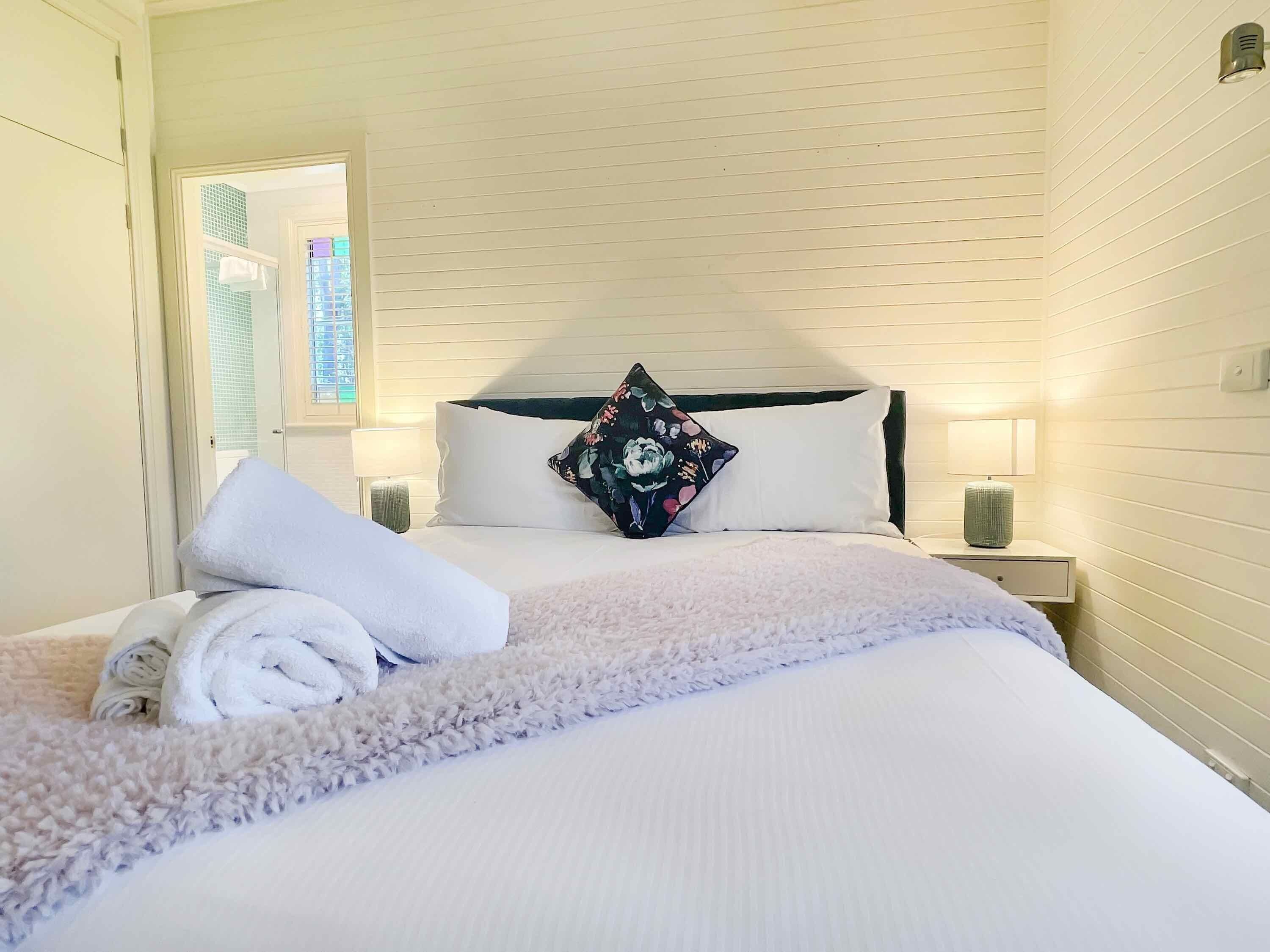 Una cama bien hecha con sábanas blancas y un tapete mullido, un cojín floral y dos lámparas de noche en una pared blanca con líneas horizontales.