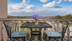 A balcony with a table and two chairs, a vase with purple flowers, and a scenic view of hills and a clear sky.