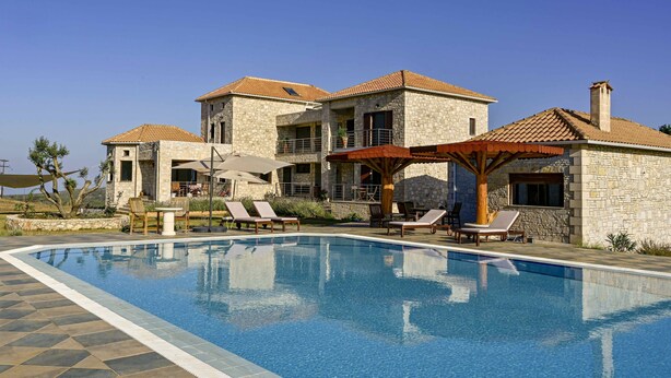 A pool area with lounge chairs and umbrellas, surrounded by stone buildings with tiled roofs.