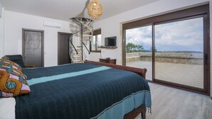 A bedroom with a bed, a chair, a TV, and a large window with a view of a stone wall and greenery.