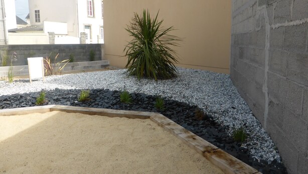 Un petit espace extérieur aménagé en jardin, avec un coin sablonneux, du gravier et un palmier.