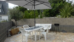 Une terrasse extérieure avec un sol en pierre, des chaises en plastique blanches, une table et un grand parasol.