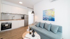 Eine moderne Küche mit weißen Schränken, einer Marmor-Spritzschutzleiste und Edelstahlgeräten. Ein graues Sofa mit Kissen, ein runder Couchtisch mit Weinflasche und Kerze, sowie ein farbenfrohes abstraktes Gemälde an der Wand.