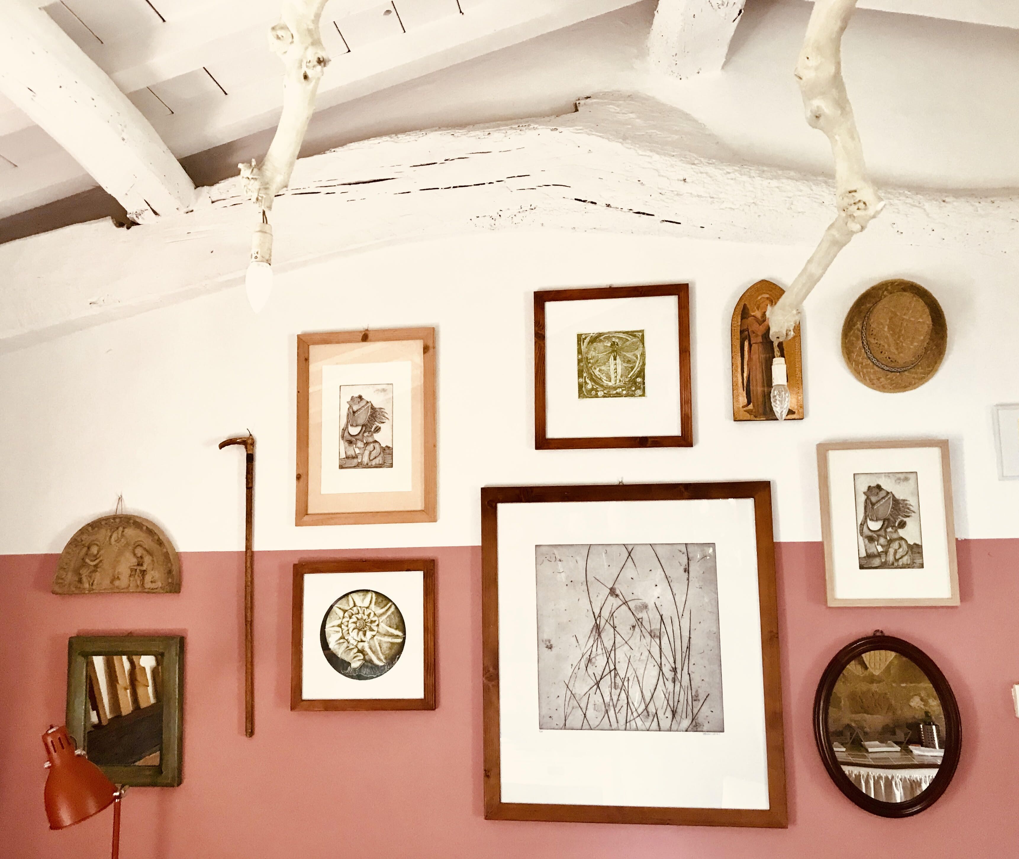 A room with a pink wall and white ceiling beams, featuring various framed artworks, a mirror, and a red desk lamp.