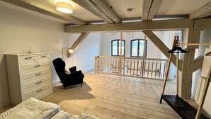 Ein Schlafzimmer mit Holzboden, einem Bett, einer Kommode, einem Stuhl und Blick ins Freie durch große Fenster.