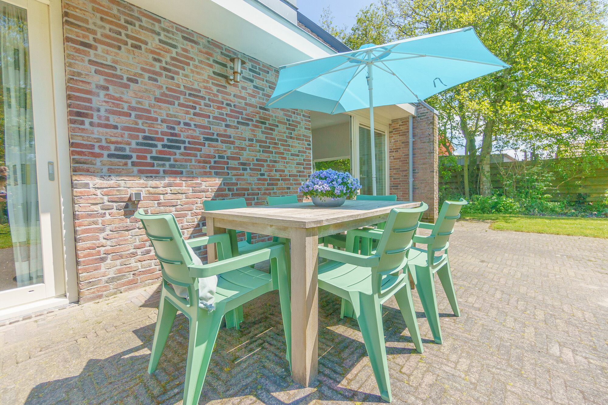 A patio with a wooden table and green chairs, a blue umbrella, and a flower pot.