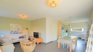 A modern living room with a sofa, a TV, a dining table with chairs, and a wicker chair. - Modern holiday home with a large garden.  Near the beach (Callantsoog)