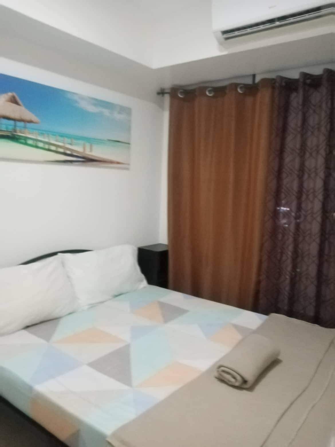 A hotel room with a bed, a painting of a beach scene, a window with brown curtains, and an air conditioning unit.