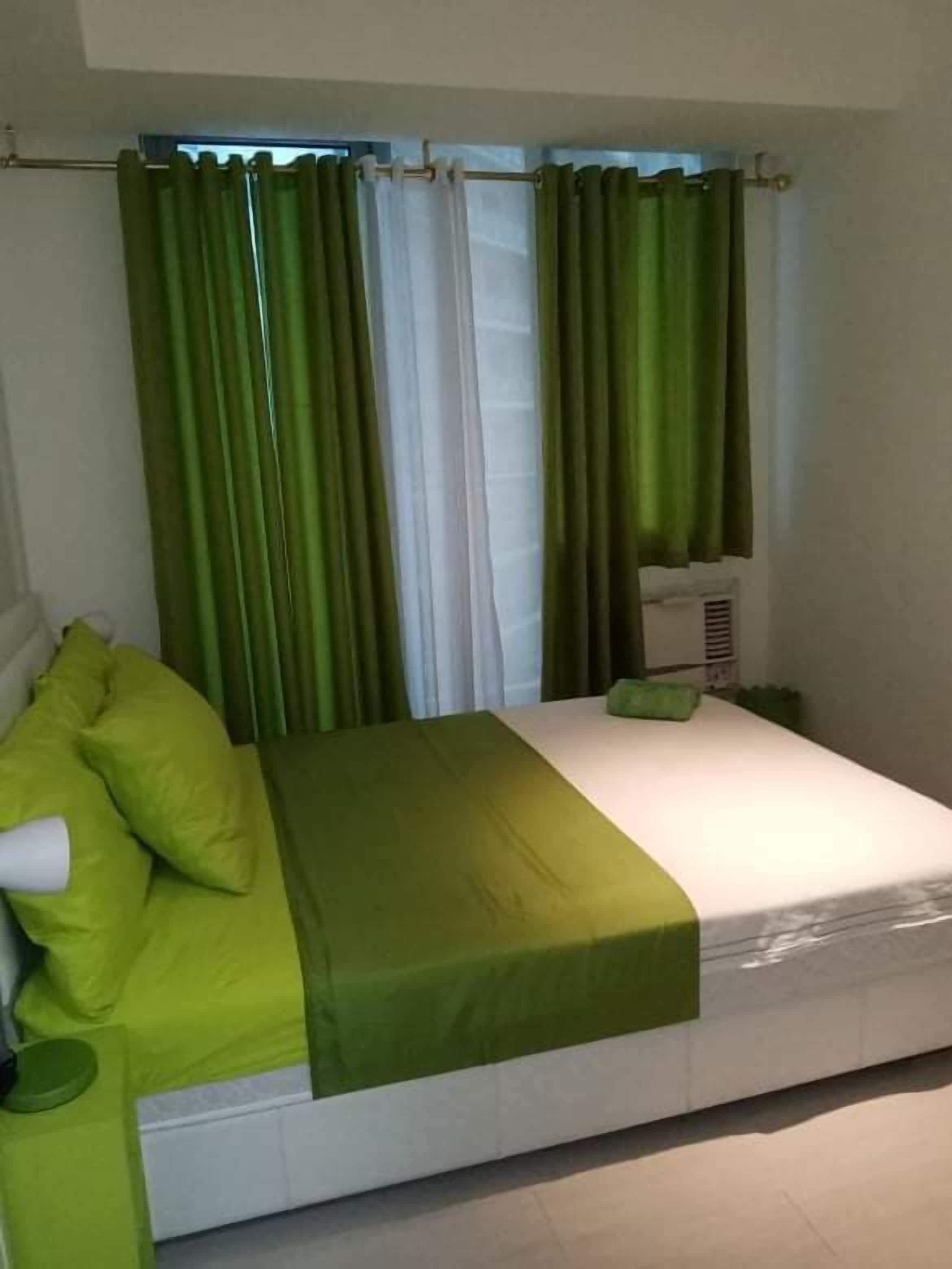 A single bed with a green bedspread and pillows, a bedside table with a lamp, and a window with green curtains.