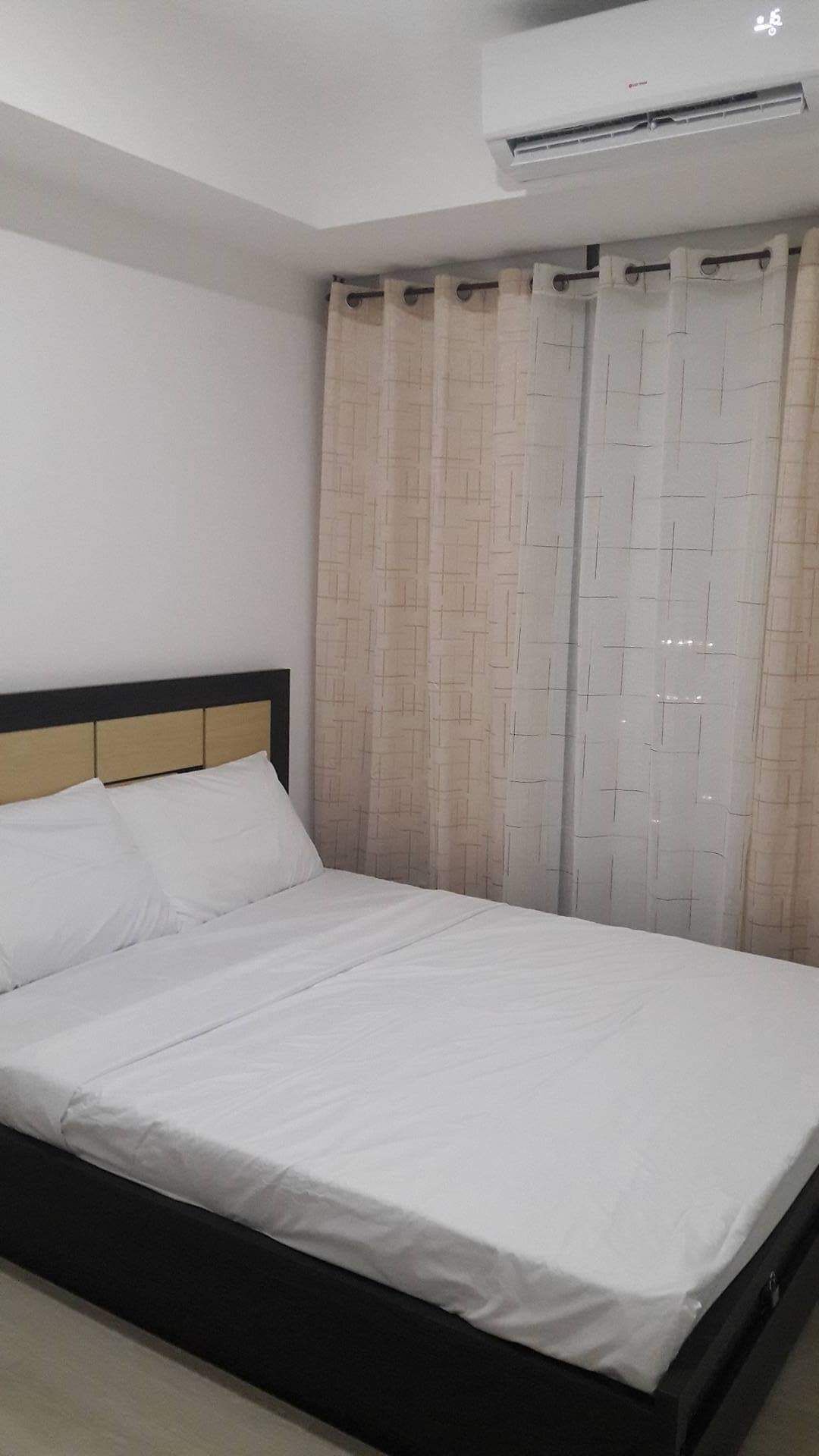 Un lit simple avec une tête de lit en bois, une literie blanche et une fenêtre avec des rideaux de couleur beige.