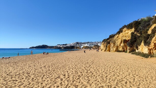 A wide sandy beach with a clear blue sky, a rocky cliff on the right, and a town in the background. - Albufeira Modern Sea View Home 33 (Albufeira)
