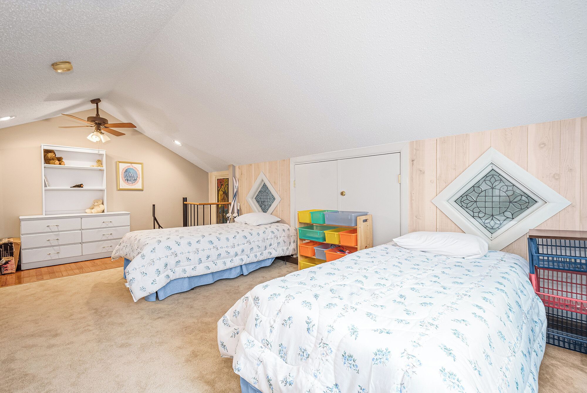 Une chambre avec deux lits, une armoire, un ventilateur de plafond et une fenêtre ornée d’un panneau de verre décoratif.