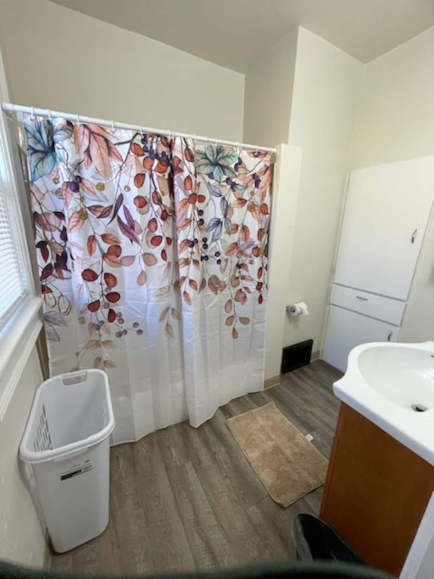 Ein Badezimmer mit Duschvorhang mit Blättermuster, einem weißen Waschbecken, einem Holzschrank und einem Mülleimer.