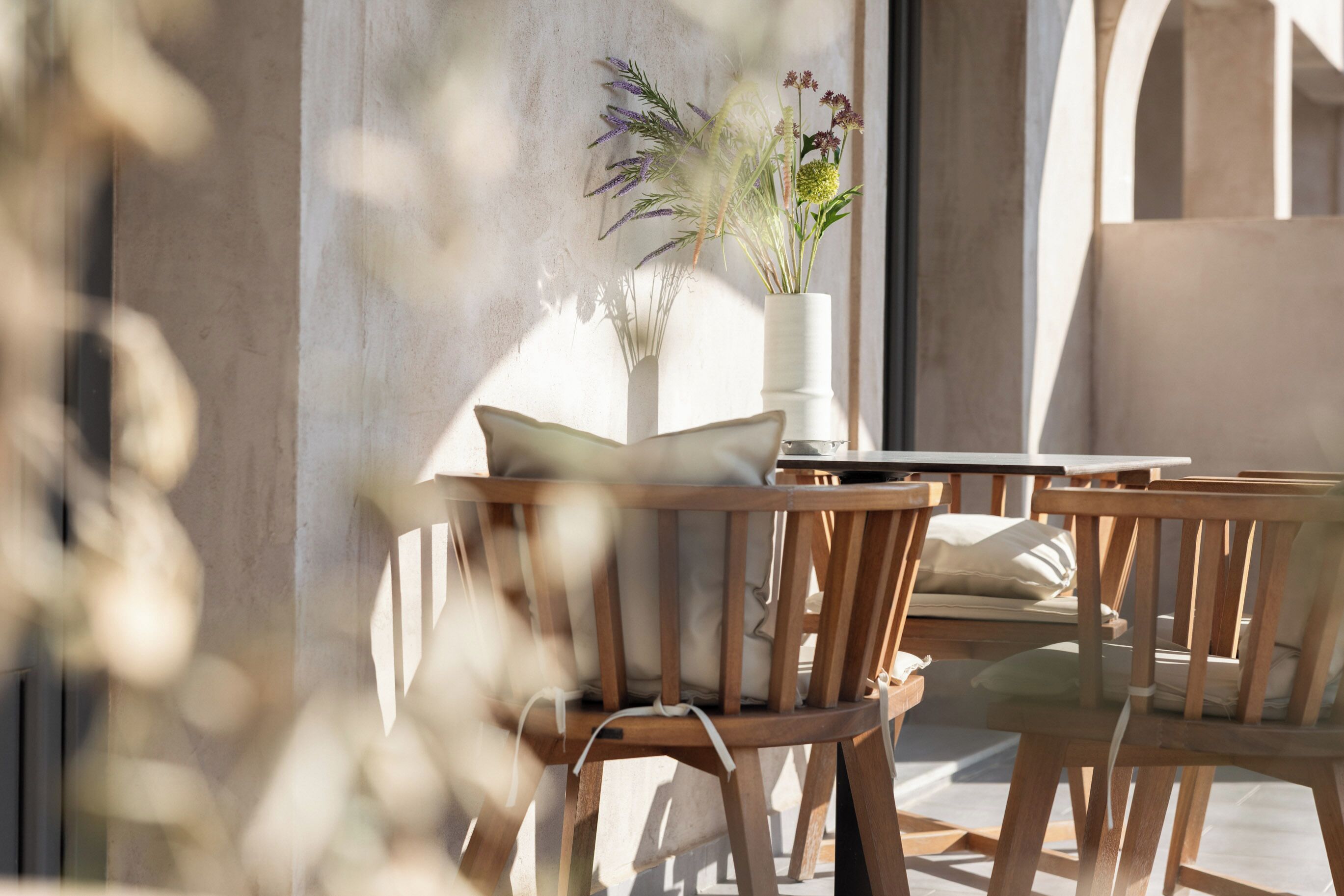 A wooden chair with a cushion, a white vase with plants, and a table in a room with a concrete wall.