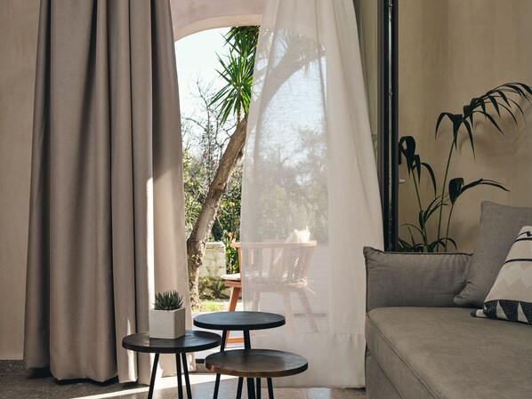 A cozy room with a sofa, a small table, and a chair. There is a window with curtains, a plant, and a view of the outdoors. - casa di pierro (Zakynthos)