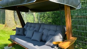 Ein modernes Outdoor-Sofa mit Holzrahmen und grauen Kissen, platziert auf einer Rasenfläche vor einem Zaun aus Metall.