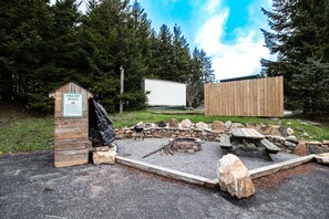 Et bålsted med en trækonstruktion, hvor der står "Fire Pit Rules", og en hvid autocamper parkeret i nærheden.