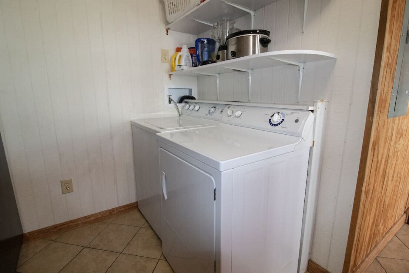 Een kleine wasruimte met een voorlader wasmachine en droger, een plank boven de droger met verschillende spullen, en een betegelde vloer.