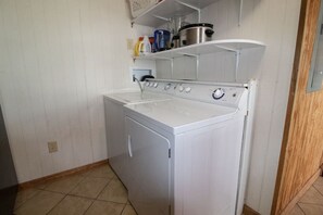 Uma lavanderia compacta com máquina de lavar e secadora frontais, uma prateleira acima da secadora com diversos objetos, e piso revestido de azulejos.