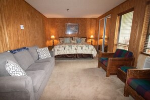 Un dormitorio con una cama, un sofá gris, un sillón colorido y un ventanal con persianas.