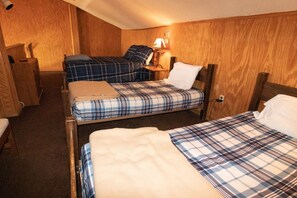 A small, cozy room with two beds, each with a plaid blanket and pillows. There is a wooden headboard, a bedside table, and a lamp on the wall.