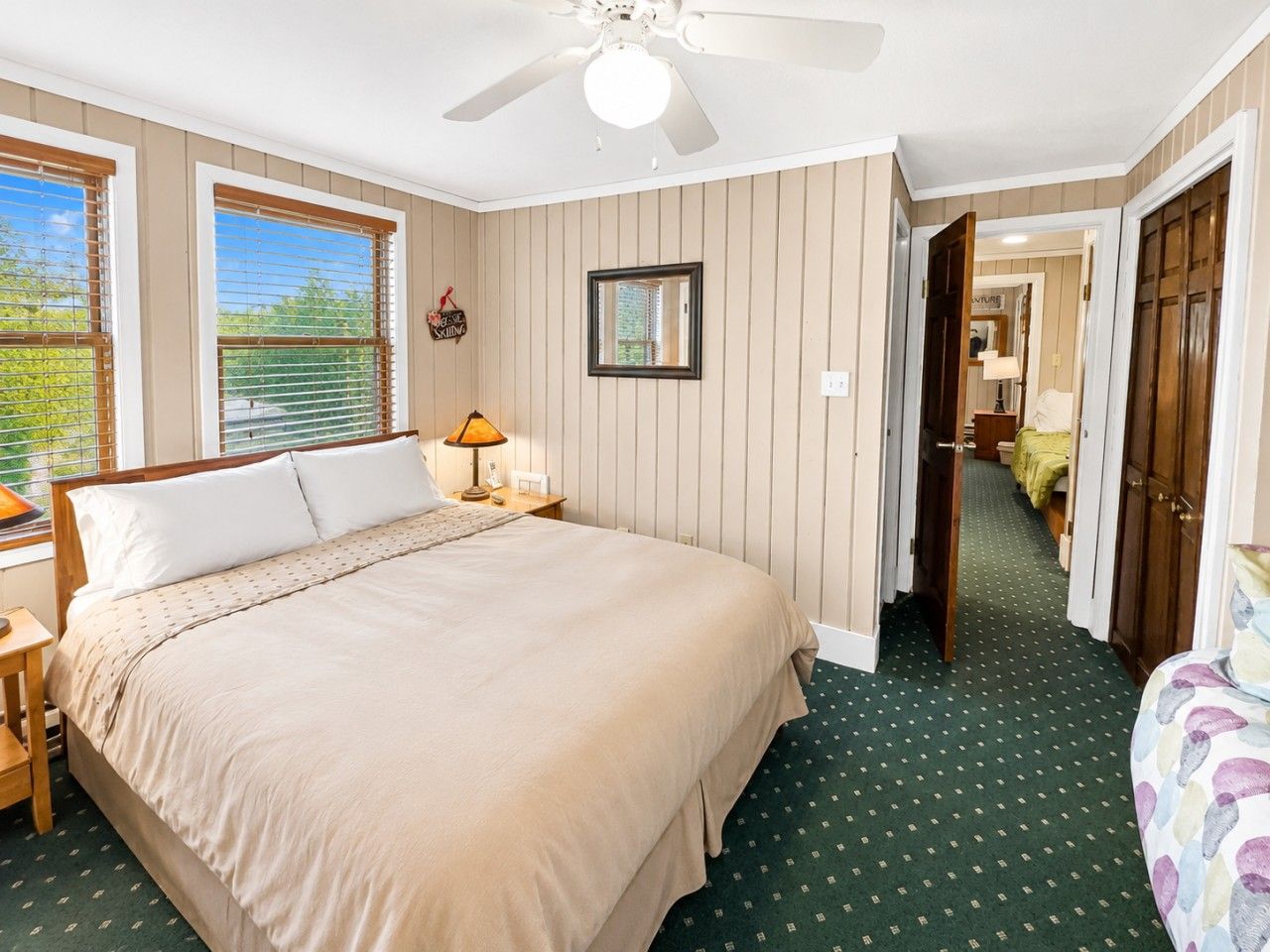 A bedroom with a bed, a nightstand, a lamp, a ceiling fan, and a view of the outdoors through the window.