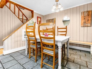 Un comedor con sillas de madera, una mesa blanca y un banco. Se observa una escalera con barandillas de madera y un candelabro.